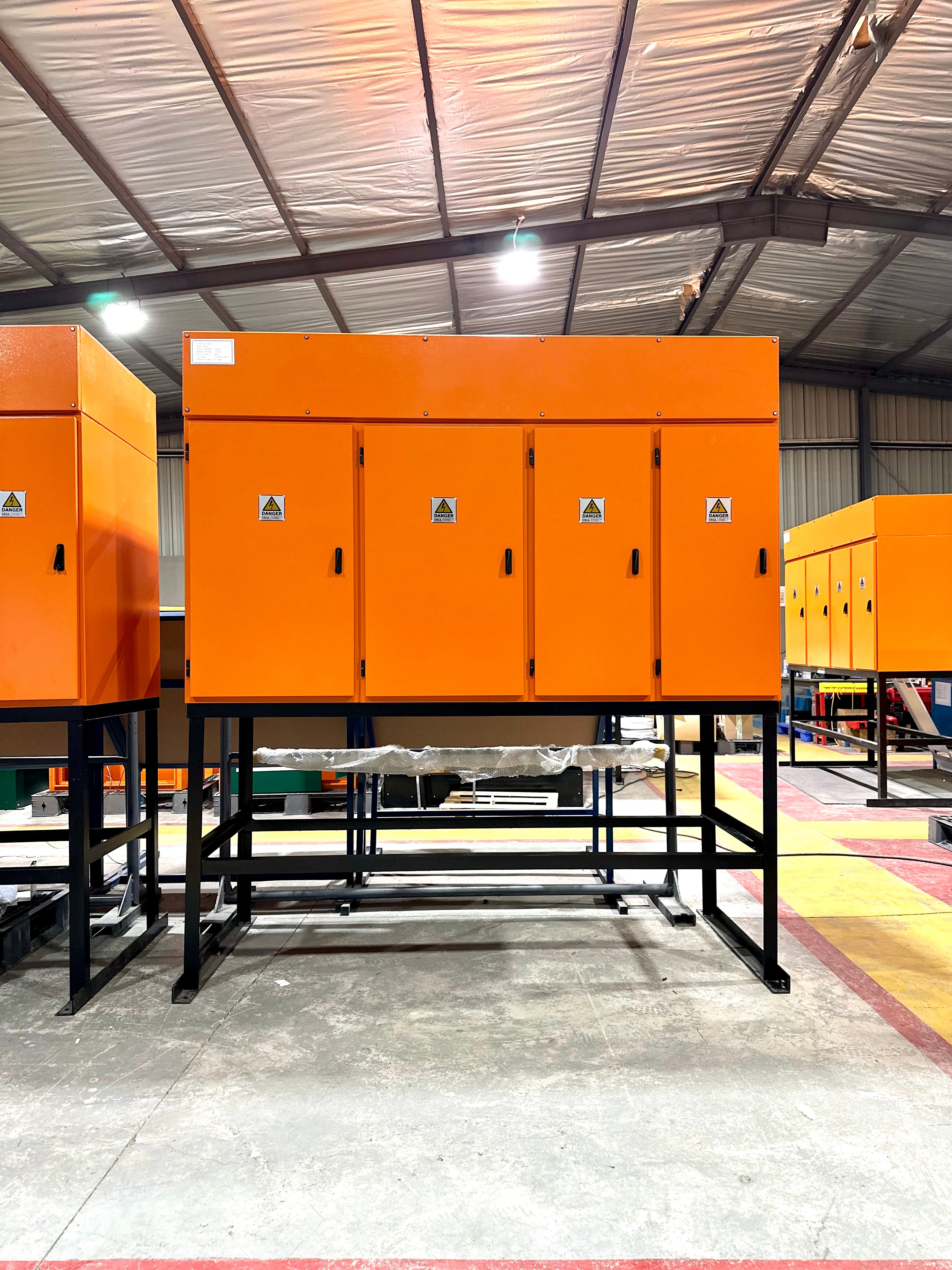 A large orange industrial electrical cabinet system mounted on a black metal frame, photographed inside a warehouse or factory with other similar cabinets visible in the background.